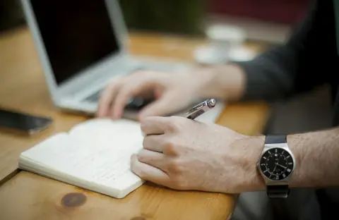 A man writing notes in a notebook next to his laptop
